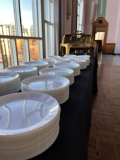 A row of white plates neatly arranged on a table with a black tablecloth, with a piano and large windows in the background.